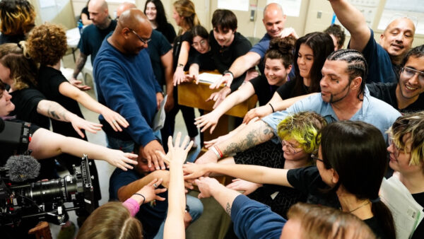 A large, diverse group stands in a circle reaching their hands into the center for a unified gesture, conveying teamwork, solidarity, and collective spirit. Some people wear blue shirts or have visible tattoos, and a professional video camera records the moment, highlighting a significant, energetic gathering or team event.