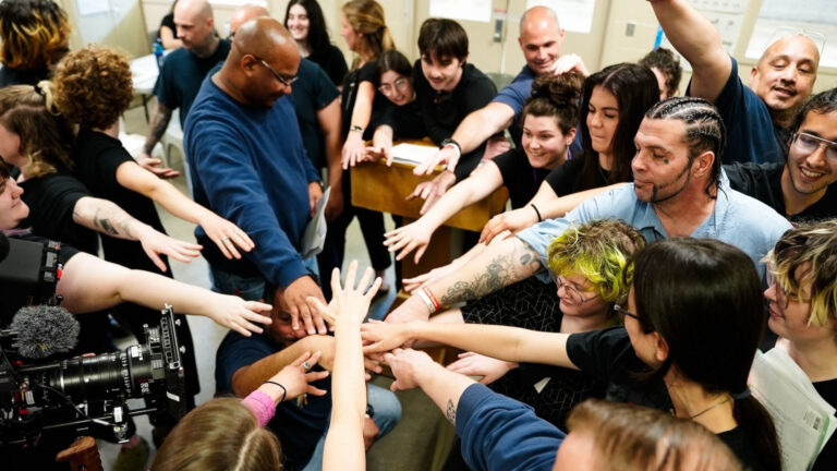 A large, diverse group stands in a circle reaching their hands into the center for a unified gesture, conveying teamwork, solidarity, and collective spirit. Some people wear blue shirts or have visible tattoos, and a professional video camera records the moment, highlighting a significant, energetic gathering or team event.