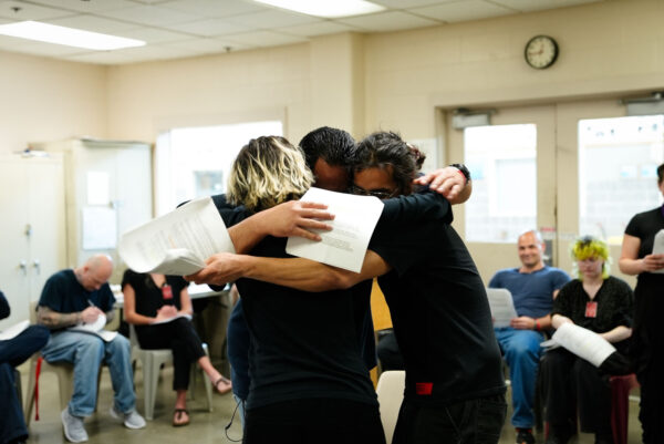 Three people stand closely in a group hug, each holding several sheets of paper, within a classroom or meeting space. Others in the background are seated and engaged in writing or reading, creating a supportive and emotionally expressive moment that suggests camaraderie, encouragement, or a shared milestone in a group setting.