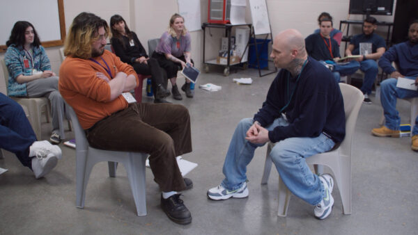 Two people sit facing each other in the center of a room, engaged in a focused and likely structured conversation, while others sit in a circle around them observing. The environment appears to be a classroom or workshop setting, with plastic chairs, casual attire, and scattered papers and notebooks, suggesting group discussion or a therapeutic or educational exercise.