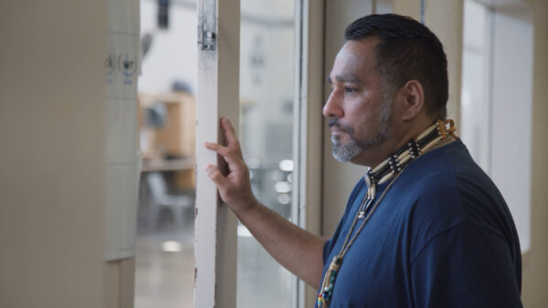 A person in a blue shirt stands indoors looking out through a window, resting one hand on the frame. They wear a necklace with several rows of beads and bone pieces, and the background is softly focused, suggesting a thoughtful or contemplative mood in an institutional or classroom environment.