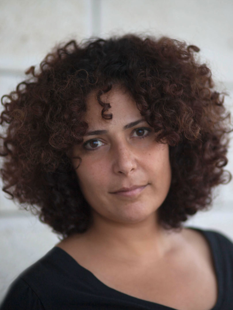 This is a portrait of a person with short, voluminous curly hair wearing a black top. The subject stands against a light-colored, softly blurred background, giving the image a clean and natural appearance.