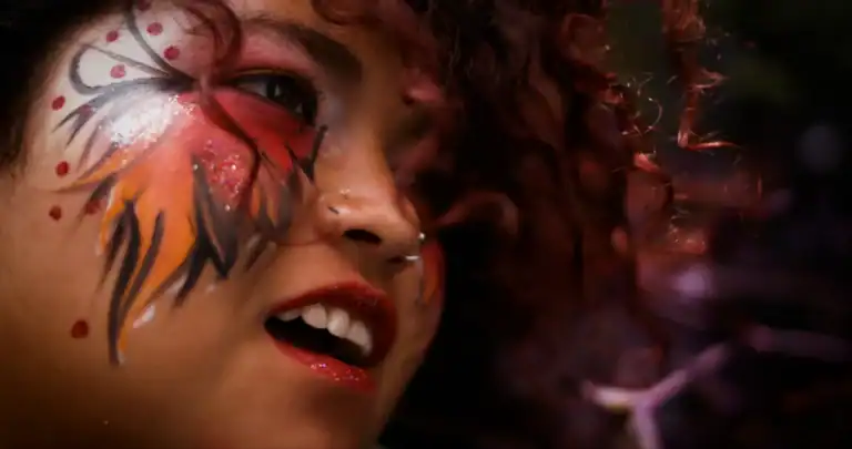 Close-up of a woman with colorful face paint, curly hair, and a nose piercing, smiling and looking to the side, with a blurred background.