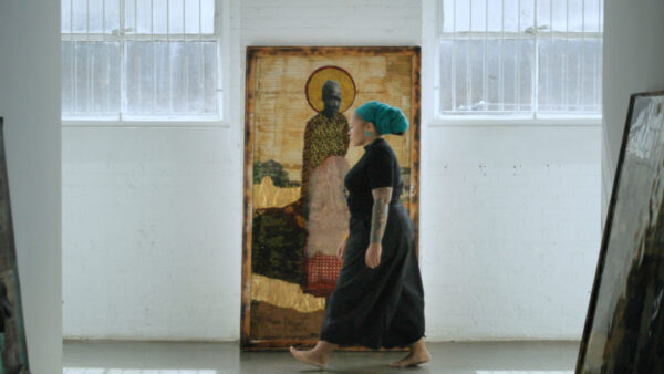 A woman wearing a turquoise headwrap, black dress, and displaying arm tattoos walks barefoot through a bright, minimalist space with white walls and barred windows. She passes a large artwork featuring a figure with a gold halo and patterned robe, evoking themes of spirituality and art within a serene, studio-like environment.