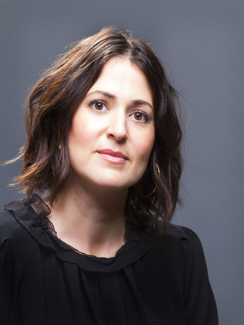 This headshot portrait features a woman with shoulder-length, softly wavy brown hair, wearing a black blouse with ruffled details and gold hoop earrings. She poses against a smooth gray background, offering a warm and professional presentation suitable for a director or creative professional.