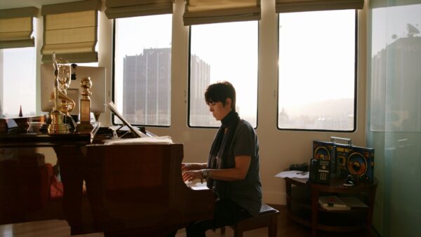 Person sitting at a grand piano playing music in a sunlit room with large windows and various awards on display.