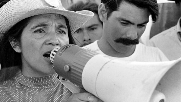 A woman wearing a hat speaks into a megaphone at a public gathering, surrounded by several people, including a man with a mustache.