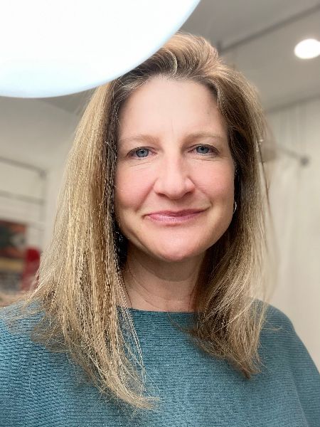 A woman with shoulder-length blonde hair and a blue sweater smiles at the camera indoors under bright lighting.