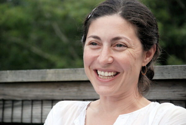 A woman with dark hair pulled back and wearing gold hoop earrings sits outdoors in front of a wooden railing, dressed in a white shirt. The background is filled with soft-focus greenery, giving the portrait a natural and relaxed atmosphere.