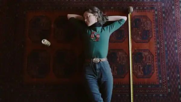 Person lying on a patterned rug with a tape measure and a small object nearby, wearing a green top and jeans, in a relaxed pose.