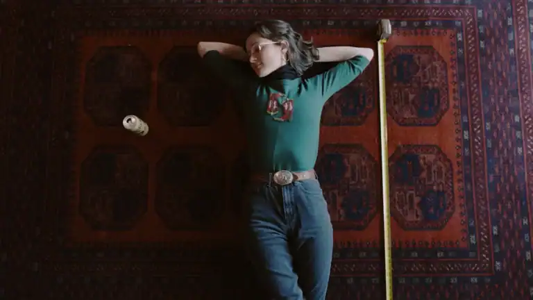 Person lying on a patterned rug with a tape measure and a small object nearby, wearing a green top and jeans, in a relaxed pose.