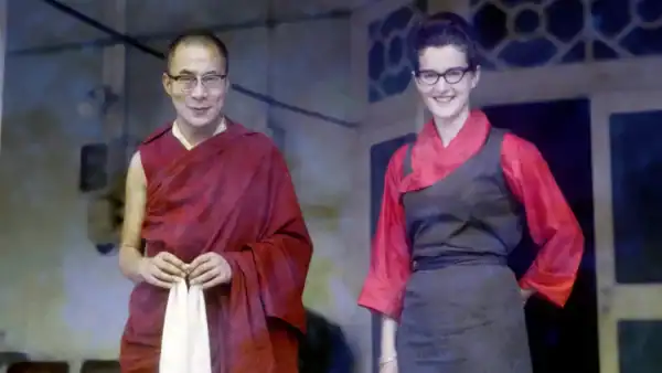 Two individuals, one in traditional Buddhist monk robes and the other in a red and black outfit, standing together and smiling at an event.