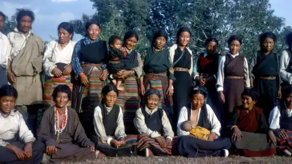Group of women and children in traditional clothing outdoors, smiling and posing for a photo in a rural setting with trees in the background.
