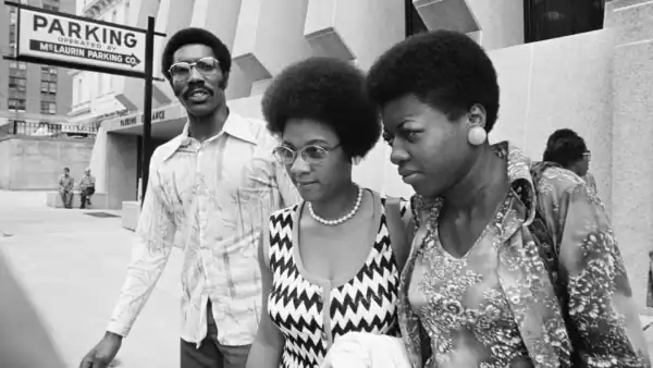 Two African American women and one man walking together on city street, engaging in conversation, with parking sign and buildings in background, black and white photo.
