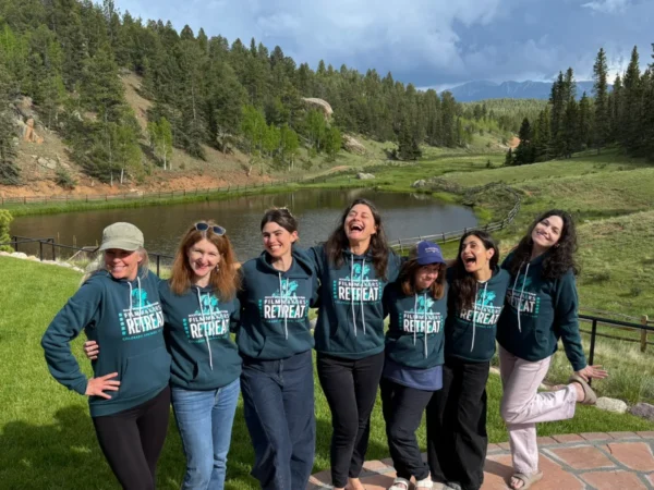 Seven women wearing matching retreat hoodies stand together smiling in front of a pond and forested hills under a partly cloudy sky.