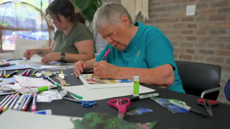 Older woman engaged in arts and crafts activity at a table with colorful markers, scissors, and paper, in a community center setting.