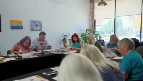 Group of diverse seniors engaging in arts and crafts activity in a bright, modern community room with large windows and colorful artwork on the walls.