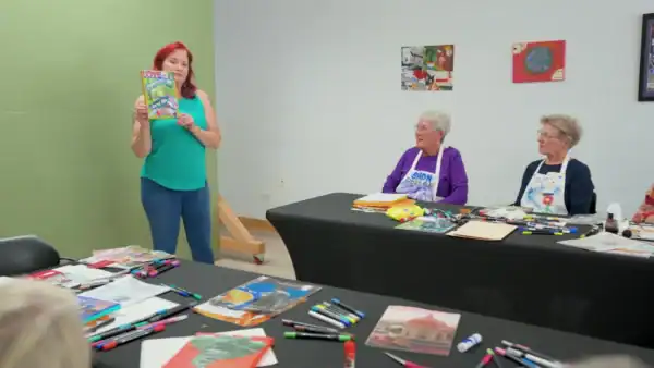 A woman in a turquoise top and blue pants presents a colorful book to two elderly women seated at a table during a community event, with art supplies and magazines on the table.