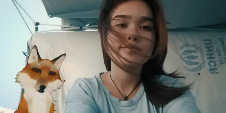 Young woman with long dark hair taking a selfie outdoors, with a fox mascot costume in the background at a film event, under a blue canopy with UNHCR logo.