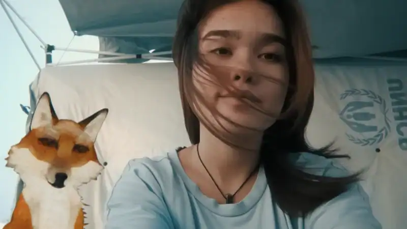Young woman with long dark hair taking a selfie outdoors, with a fox mascot costume in the background at a film event, under a blue canopy with UNHCR logo.