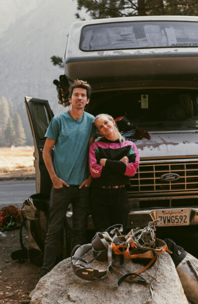 Two climbers stand in front of a rugged Ford camper van, surrounded by climbing gear and ropes. One leans with arms crossed and a relaxed expression, while the other stands casually with hands in pockets. A large pile of harnesses and equipment sits on a boulder in the foreground, capturing the laid-back yet adventurous atmosphere typical of a big-wall climbing trip.