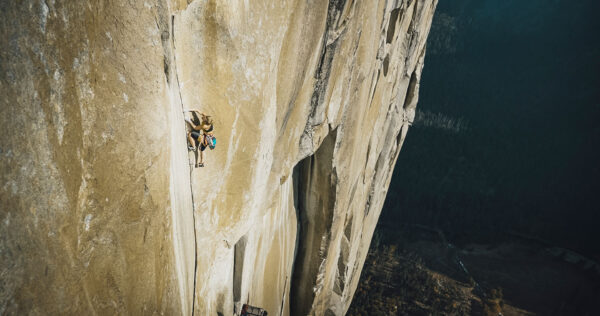 A climber ascends a massive, nearly vertical granite face, high above the valley floor. The cliff is sheer and dramatic, with the climber skillfully maneuvering up a crack system, carrying gear and rope. The vast scale and natural beauty of the rock wall emphasize the challenge and daring involved in big-wall climbing.
