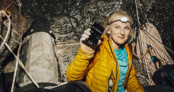 The image shows a climber in a yellow puffer jacket sitting at a rocky ledge, surrounded by climbing ropes and equipment, holding up a smartphone with a stopwatch timer reading over 21 hours. The climber is in a rugged outdoor setting, conveying endurance and determination likely during a multi-day climb or expedition.