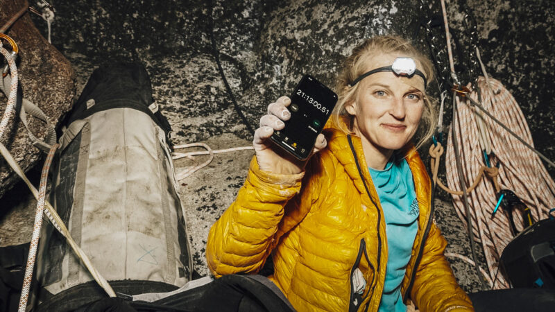 The image shows a climber in a yellow puffer jacket sitting at a rocky ledge, surrounded by climbing ropes and equipment, holding up a smartphone with a stopwatch timer reading over 21 hours. The climber is in a rugged outdoor setting, conveying endurance and determination likely during a multi-day climb or expedition.