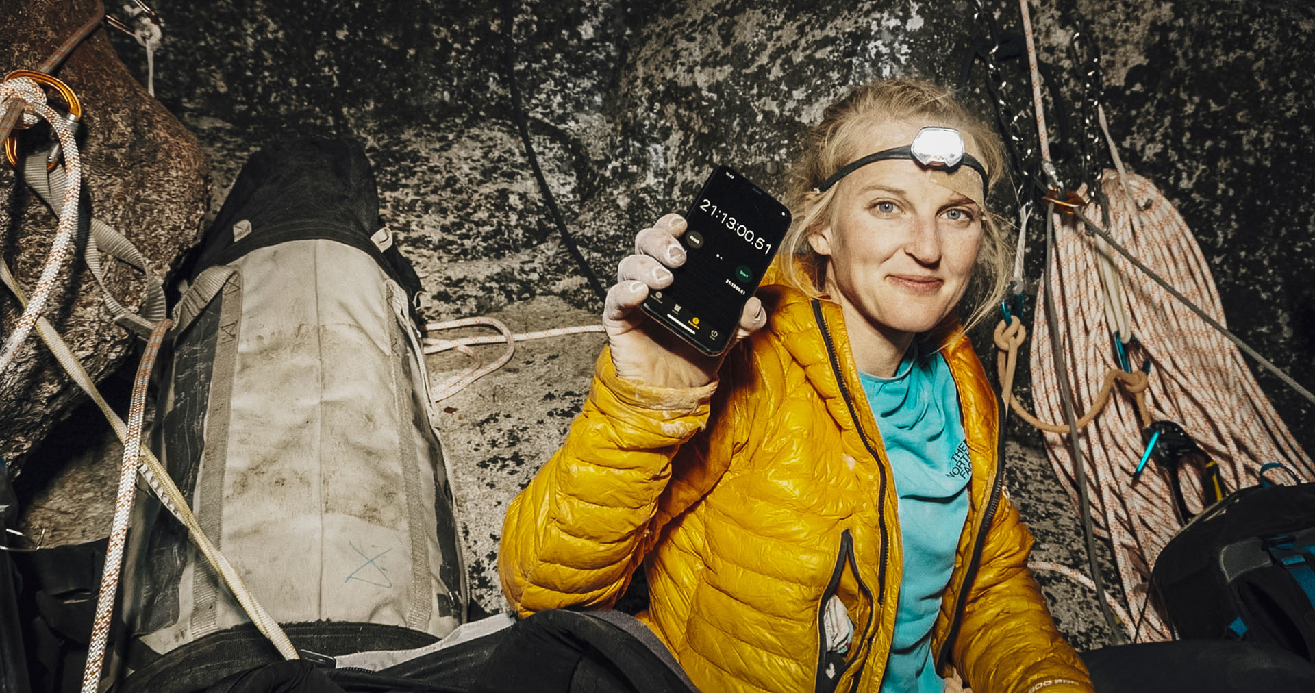 The image shows a climber in a yellow puffer jacket sitting at a rocky ledge, surrounded by climbing ropes and equipment, holding up a smartphone with a stopwatch timer reading over 21 hours. The climber is in a rugged outdoor setting, conveying endurance and determination likely during a multi-day climb or expedition.