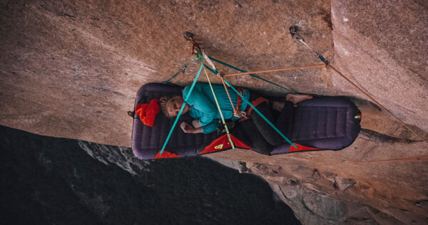 A climber is resting on a portaledge—a portable platform anchored to the side of a steep cliff. The person is bundled in outdoor gear, lying flat and secured by multiple harnesses and ropes, with dramatic rock and open air below, exemplifying the unique realities of multi-day big wall climbing expeditions.