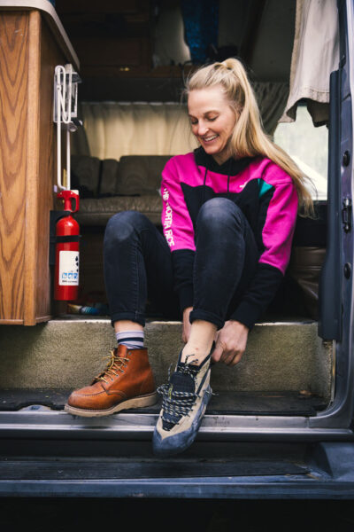 A climber sits on the step of a van interior, smiling while changing footwear—one foot already in a climbing shoe, the other still in a brown boot. The scene, complete with a visible fire extinguisher, highlights the comfortable, everyday rituals and preparations of life on the road for a dedicated climber living in a van.