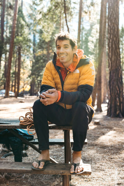 A climber dressed in a yellow and gray jacket and black pants sits relaxed on a picnic table bench in a pine forest campsite. Wearing sandals, with climbing rope coiled nearby, the person looks ready for adventure or taking a break in the fresh air of an outdoor climbing destination.