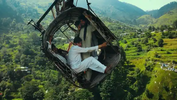 People in a damaged metal cable car suspended high above a green, hilly landscape.