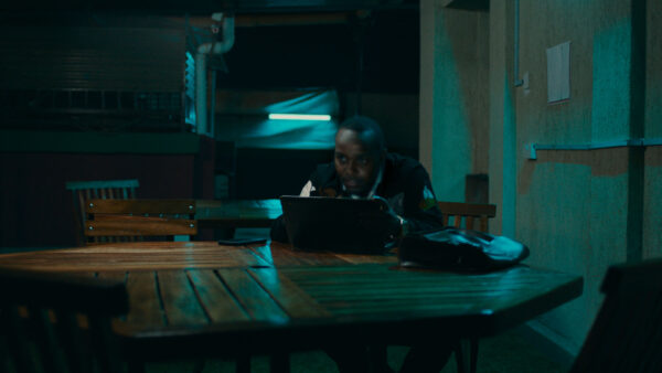 A person sits alone at a wooden table in a dimly lit room, focused on a laptop.