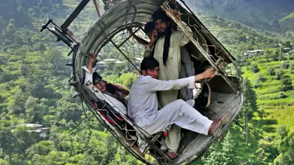 People in a damaged metal cable car suspended high above a green, hilly landscape.