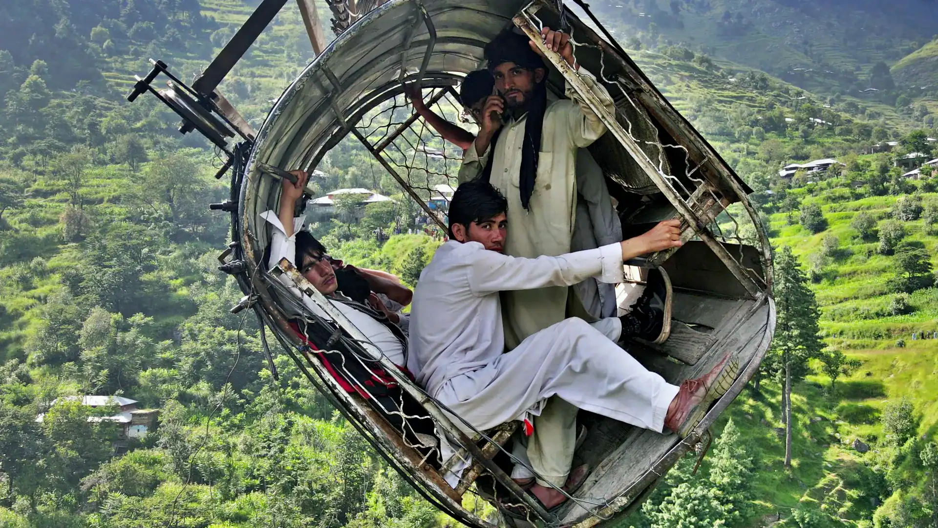 People in a damaged metal cable car suspended high above a green, hilly landscape.