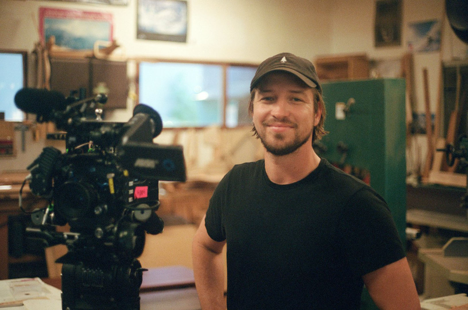 A person stands in a well-lit workshop or studio space next to a professional video camera mounted on a tripod. The individual wears a dark t-shirt and a baseball cap, with various tools and artwork visible in the background, indicating a creative production environment.