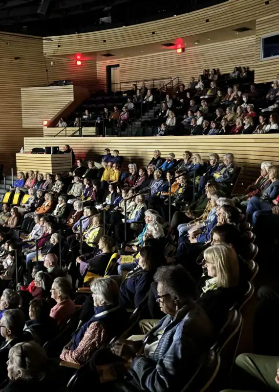 An audience seated in a tiered auditorium with wooden slatted walls.