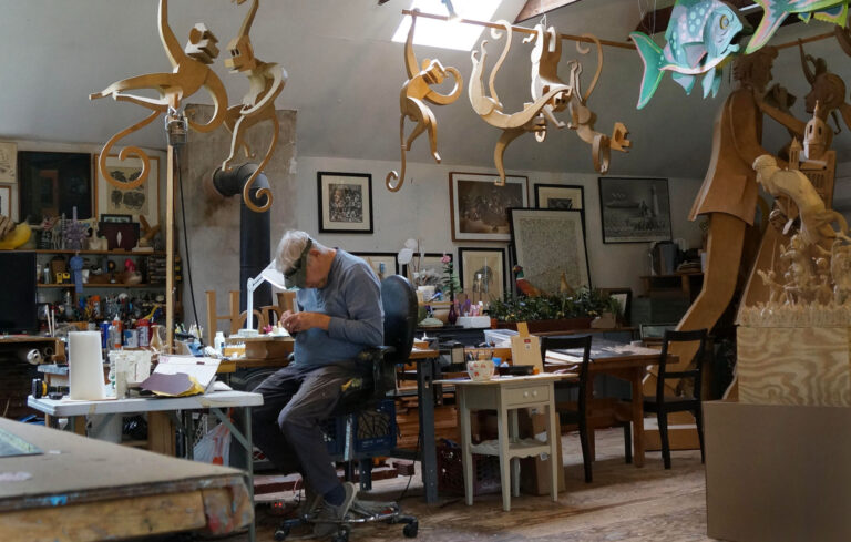 A man works at a cluttered desk in an art studio filled with hanging wooden animal sculptures and various art supplies.