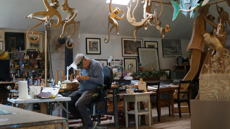A man works at a cluttered desk in an art studio filled with hanging wooden animal sculptures and various art supplies.