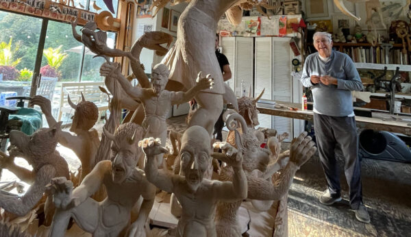 A man stands in a sunlit studio filled with large, expressive wooden sculptures of various human and fantastical figures.