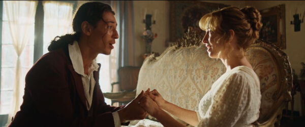 A man and woman in period clothing sit on an ornate sofa, holding hands. Sunlight filters through a window, creating a warm, intimate atmosphere.
