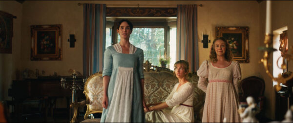 Three women in regency-era dresses stand and sit in an elegant living room with vintage furniture and framed art, conveying a period drama atmosphere.