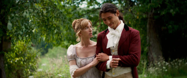 A woman in a Regency-style dress holds a man's arm tenderly. He wears a burgundy coat with a white cravat. They stand amidst lush greenery, conveying a gentle, romantic atmosphere.