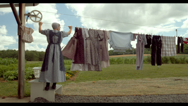A woman wearing a traditional dress and bonnet stands on a small step, hanging laundry on an outdoor clothesline. Dresses, shirts, and pants are clipped to the line, and a white laundry basket sits beside her. The grassy yard, garden, and fields stretch out under a partly cloudy sky, creating a peaceful rural scene.