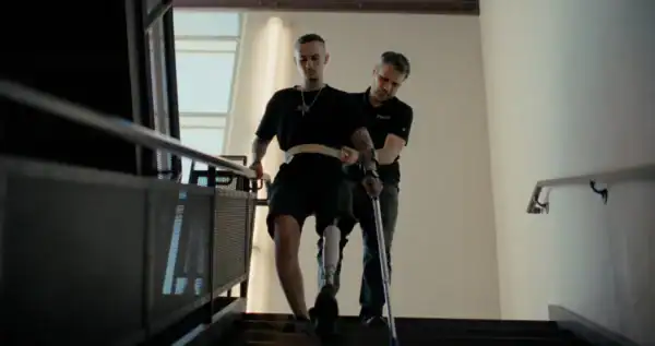 A young man with a cane being assisted by a caregiver down a staircase in a modern building with large windows and natural light.