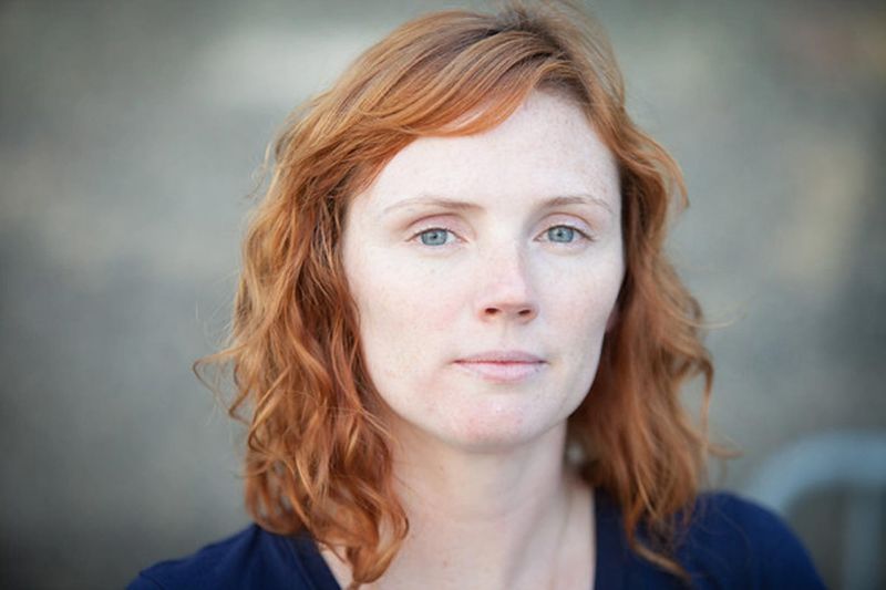 A woman with wavy red hair and light skin looks directly at the camera with a neutral expression. She is wearing a dark top and is outdoors.