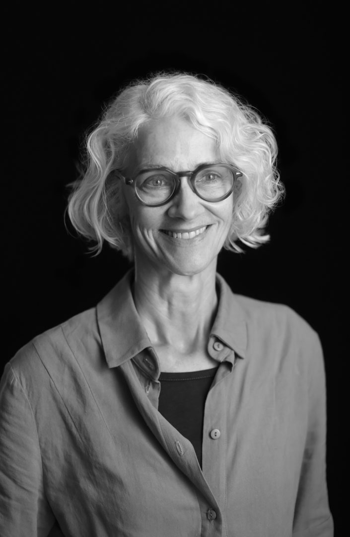 A black-and-white portrait shows a person with light, curly hair wearing an open-collared shirt over a dark top. The individual is posed in front of a plain, dark background, capturing a professional and artistic atmosphere.