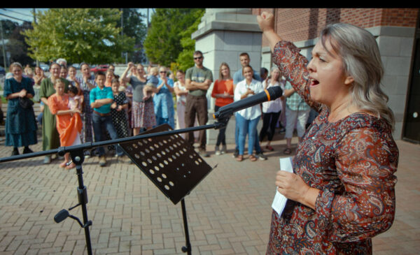 A woman with long gray hair stands at a microphone outdoors in front of a crowd, holding a piece of paper and raising her arm in a gesture of celebration or solidarity. The audience, consisting of men, women, and children in colorful dresses and casual clothing, watches attentively. The scene takes place on a paved area near a brick building and leafy tree, suggesting a public gathering or community event.