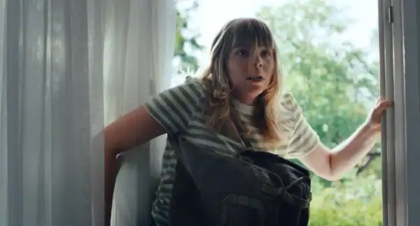 Young woman climbing in a window with a surprised expression, standing in a room with natural light and greenery outside.